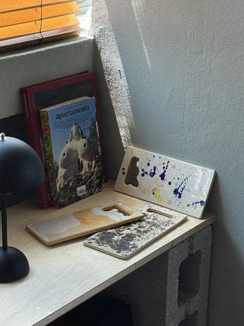 A corner desk with three Mirjam Faber Ceramics Sushi Plates with abstract cutouts, a black lamp, and a stack of magazines including Apartamento, all set by a concrete wall under a window with orange blinds.