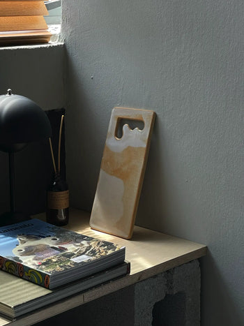 The Mirjam Faber Ceramics Sushi Plate, featuring abstract beige and white patterns, leans against a gray wall on a wooden desk by a window, beside a book, lamp, and reed diffuser.