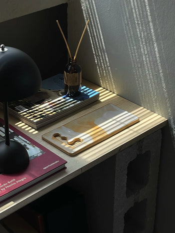A sunlit desk with a black lamp, brown glass diffuser, magazines, and a Mirjam Faber Ceramics Sushi Plate featuring unique cutouts. Sunlight casts striped shadows through blinds onto the light wood surface.