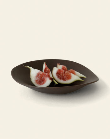 The NR Ceramics HIN Dessert Plate by NR Ceramics, in a dark modern design, displays two sliced figs with red centers and pale green skin against a simple, light background.