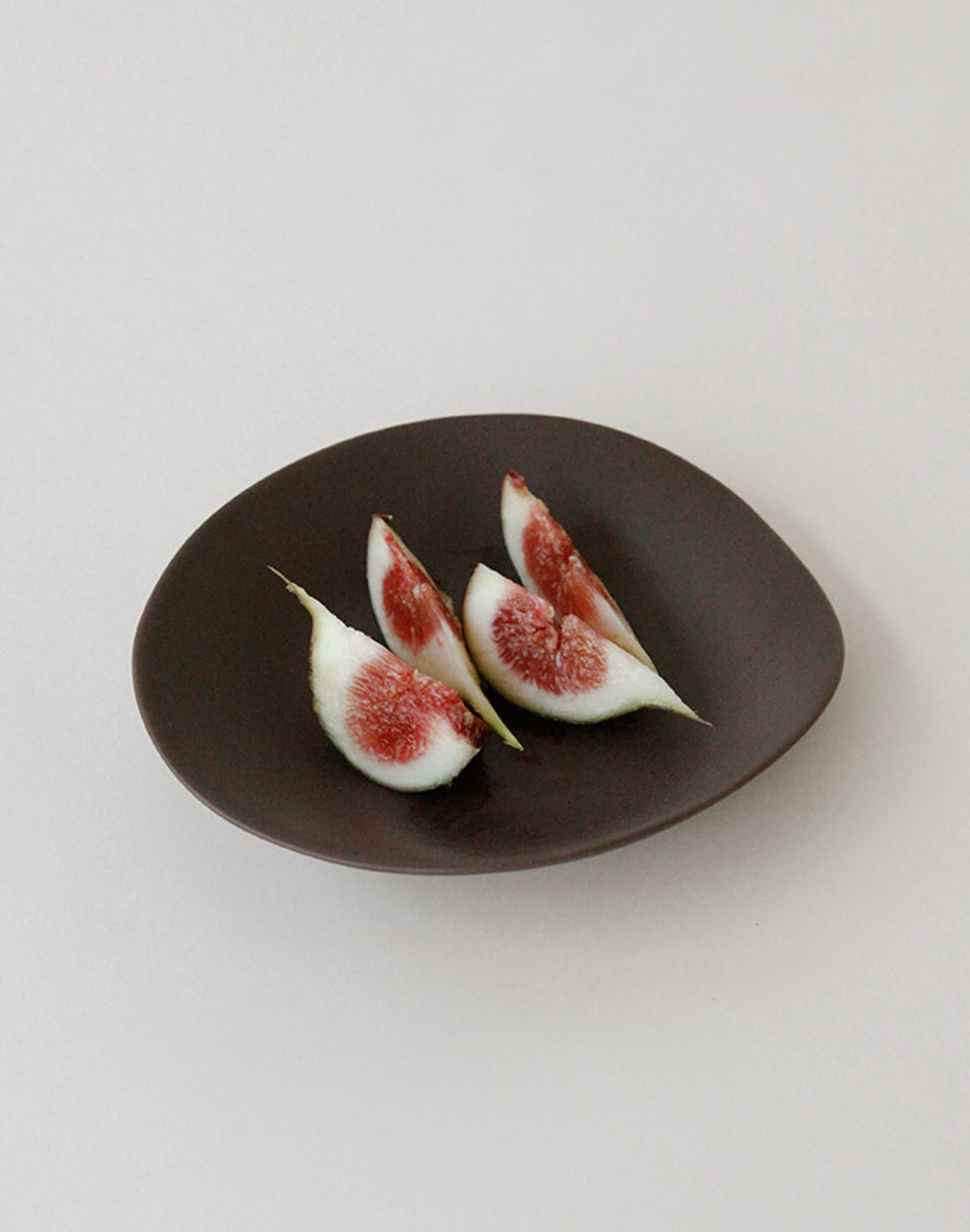 Four slices of fresh fig with red centers and white-green skin are arranged on the NR Ceramics HIN Dessert Plate by NR Ceramics, highlighting their organic shape against a simple, light background.
