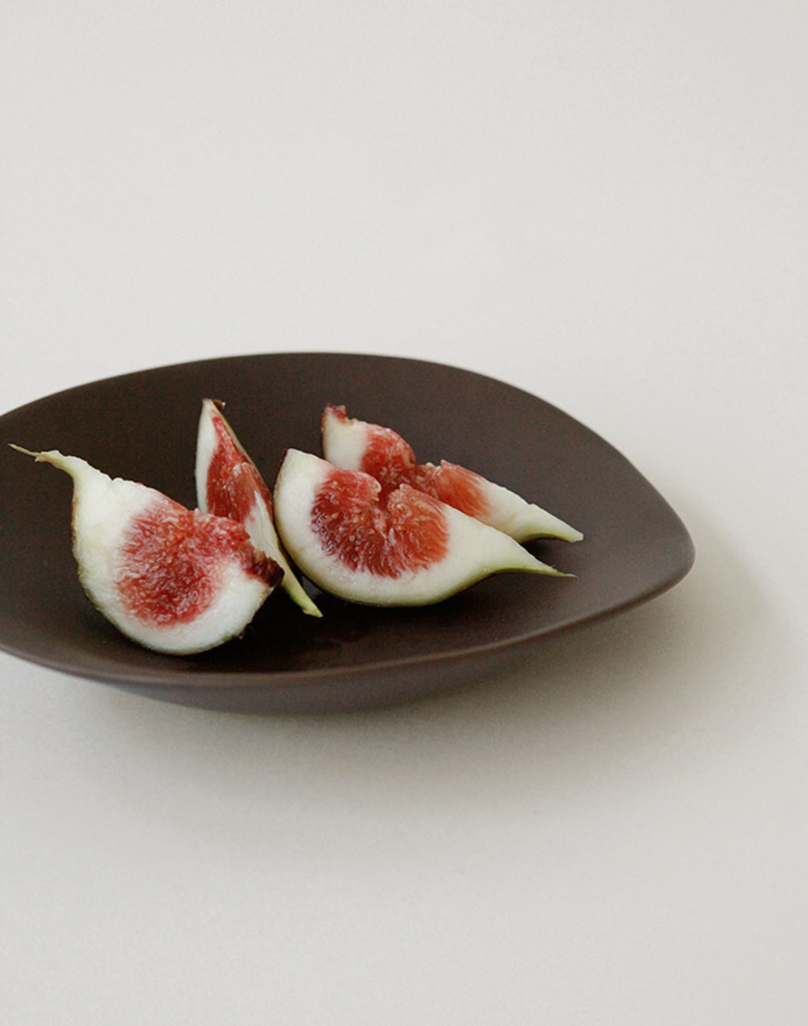 Four slices of fresh fig are arranged on the NR Ceramics HIN Dessert Plate by NR Ceramics, set against a plain white background.