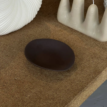 The NR Ceramics HIN Dessert Plate, with its small, dark brown, organic shape, rests on a cork surface beside a textured white vase and a cream sculptural candle.