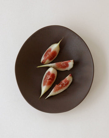 Four peeled fig quarters with red interiors are arranged in a spiral on an NR Ceramics HIN Dessert Plate, the plate’s organic shape by NR Ceramics contrasting elegantly against a plain white background.