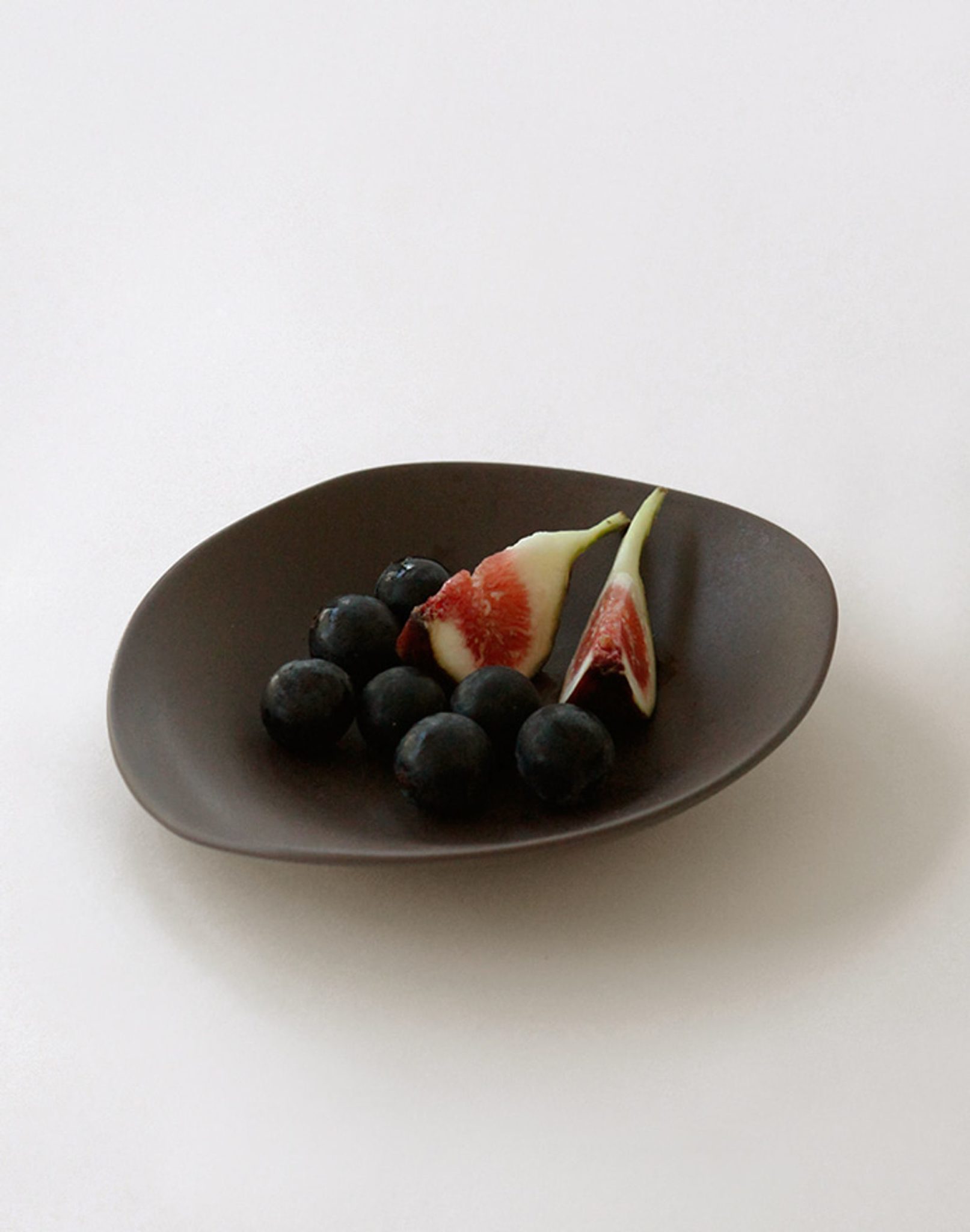 The NR Ceramics HIN Dessert Plate by NR Ceramics, featuring an organic dark design, displays black grapes and two fresh fig slices on a plain white background.