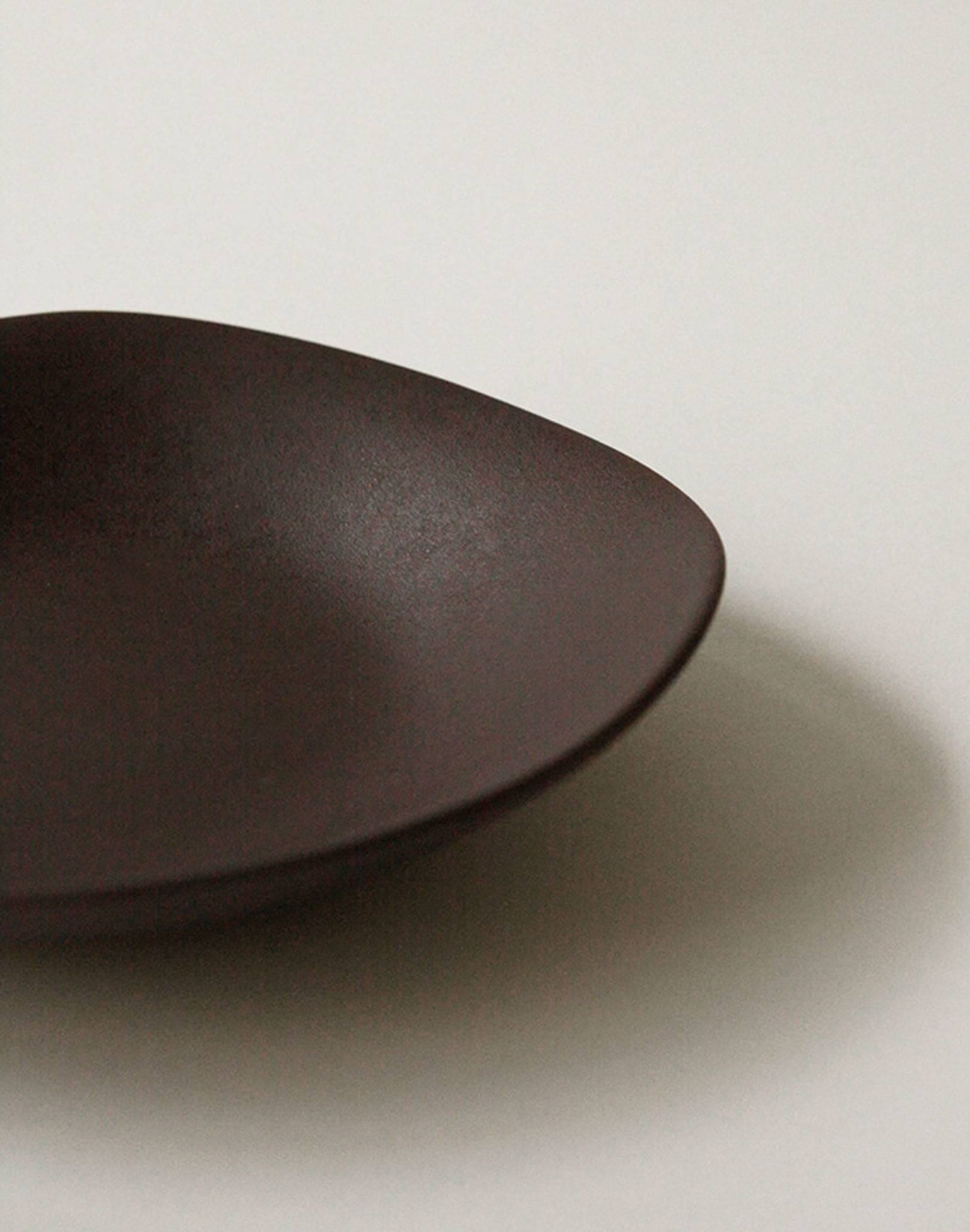A close-up of the NR Ceramics HIN Dessert Plate by NR Ceramics, featuring a dark brown modern oval shape, rests on a plain light background with a gentle shadow to the right that highlights its organic form.