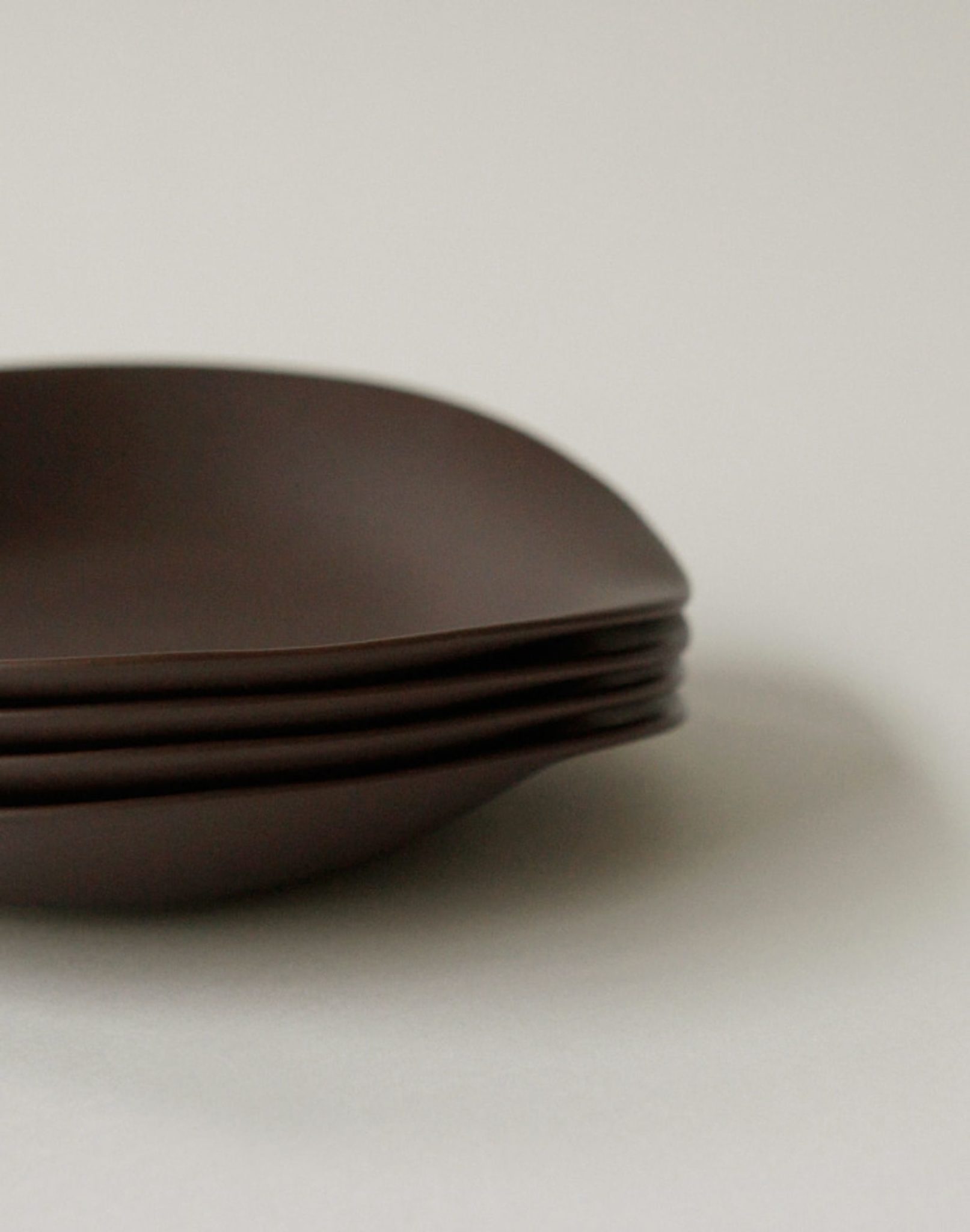 A close-up side view shows a stack of four NR Ceramics HIN Dessert Plates, each matte brown with an organic shape, against a plain light background.