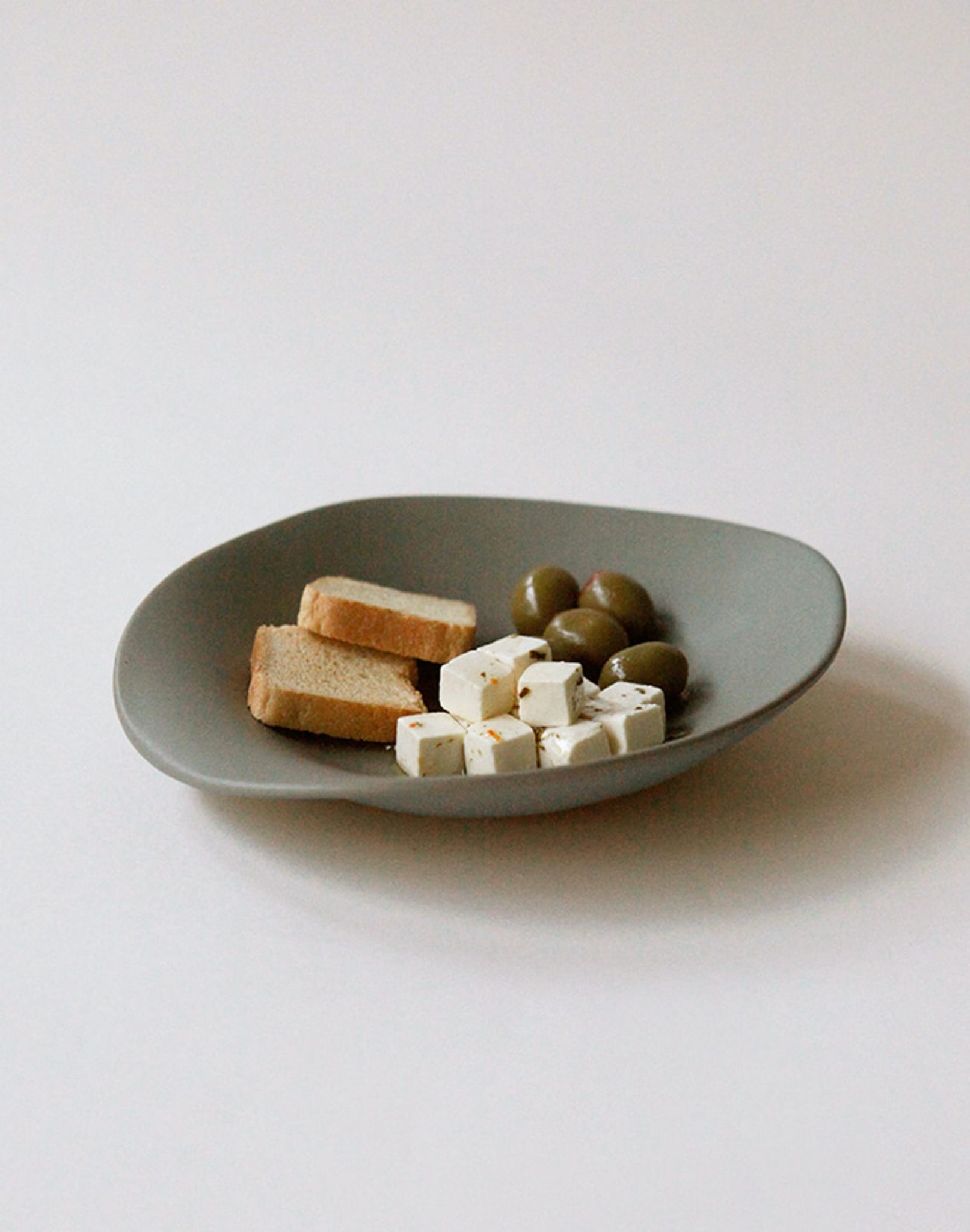 The NR Ceramics HIN Dessert Plate by NR Ceramics, in gray with an organic shape, holds green olives, white cheese cubes, and two pieces of toasted bread on a plain white background, adding modern charm to any setting.