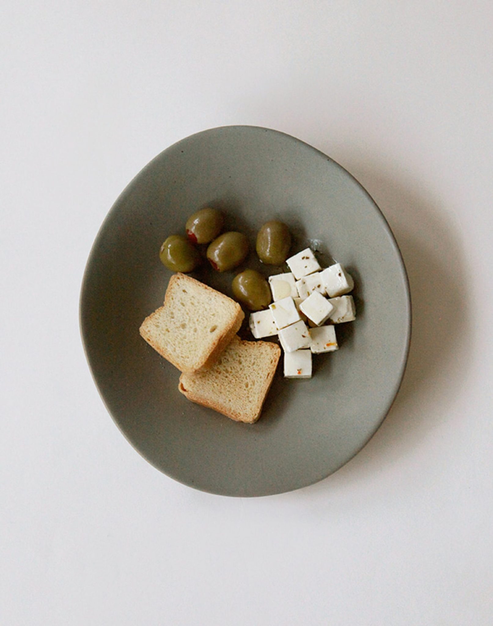 The NR Ceramics HIN Dessert Plate, in gray with an organic shape, features green olives, herbed cubed white cheese, and toasted bread slices. From NR Ceramics, this modern plate is shown against a white background.