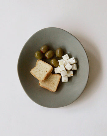 The NR Ceramics HIN Dessert Plate, in gray with an organic shape, features green olives, herbed cubed white cheese, and toasted bread slices. From NR Ceramics, this modern plate is shown against a white background.