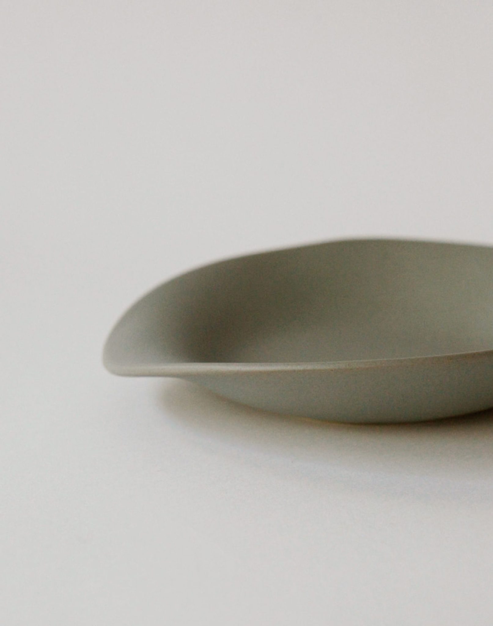 A close-up of the NR Ceramics HIN Dessert Plate, a minimalist matte gray piece by NR Ceramics featuring smooth, curved edges and an organic shape, partially shown against a light neutral background.