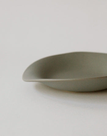 A close-up of the NR Ceramics HIN Dessert Plate, a minimalist matte gray piece by NR Ceramics featuring smooth, curved edges and an organic shape, partially shown against a light neutral background.