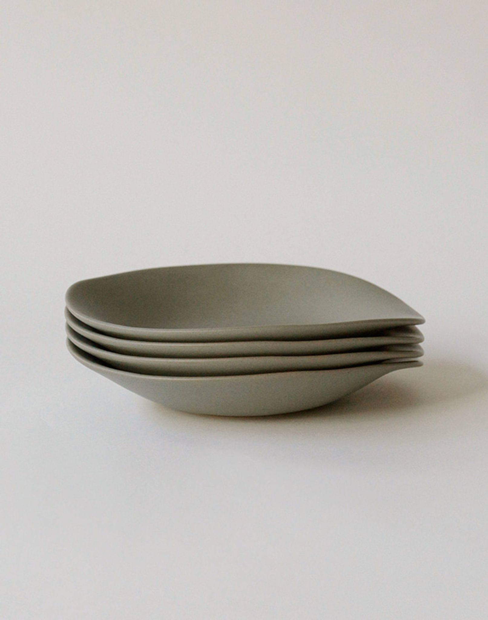 A stack of four NR Ceramics HIN Dessert Plates—minimalist, matte grey pieces with curved, organic shapes by NR Ceramics—rests on a plain white surface.