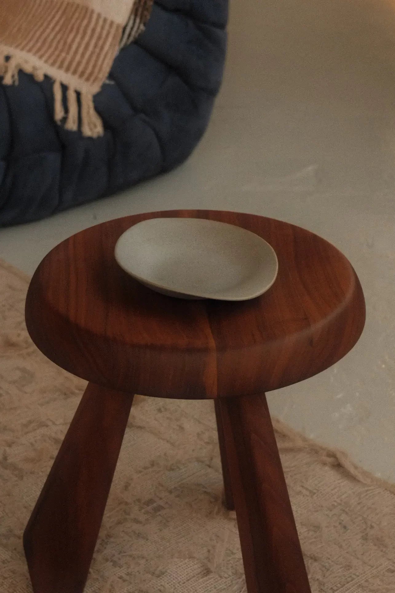 A small, round wooden stool with three legs holds the NR Ceramics HIN Dessert Plate. A textured rug lies underneath, while a blue cushioned seat draped with a fringe blanket is visible in the background.