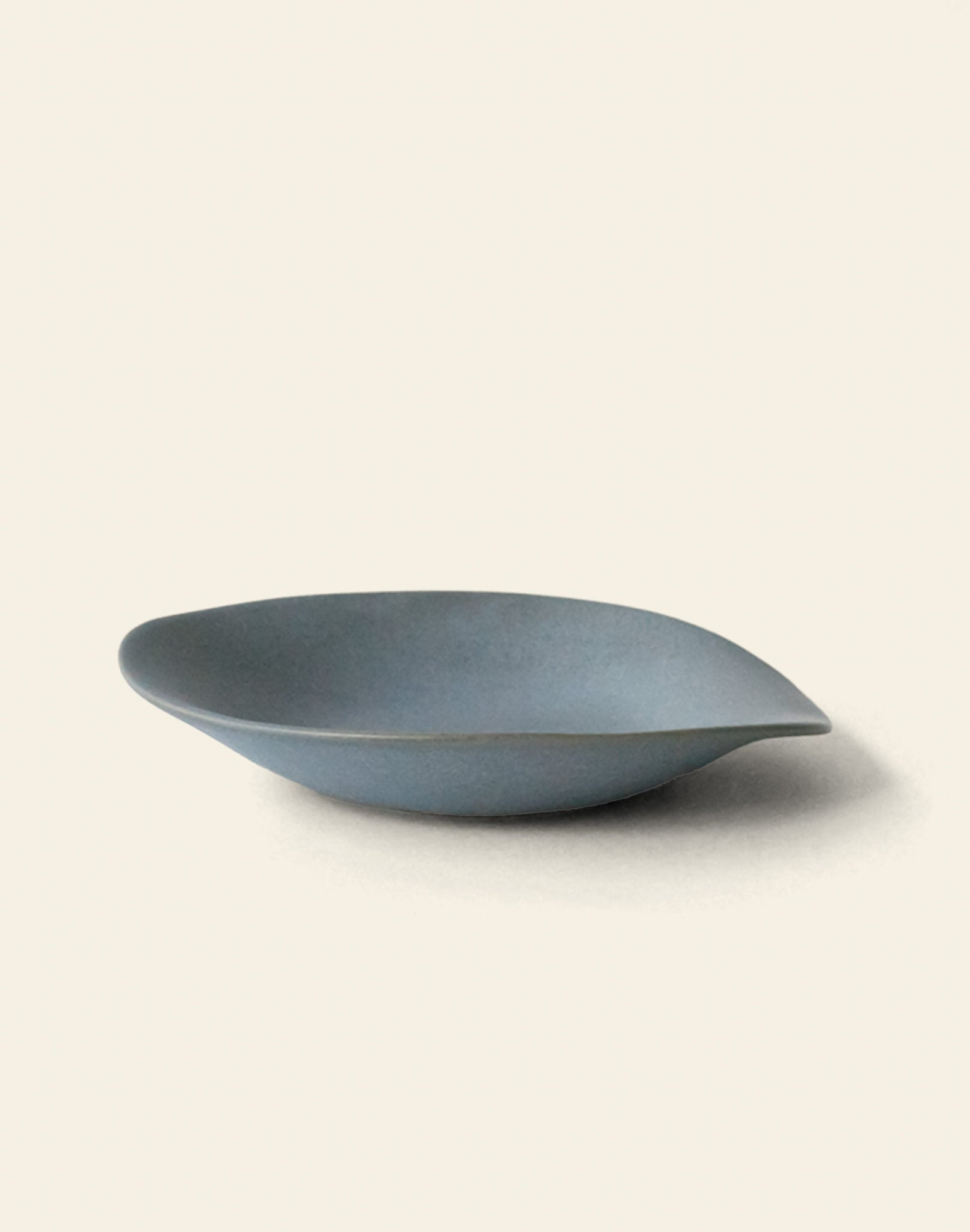 The NR Ceramics HIN Dessert Plate by NR Ceramics is a minimalist, matte blue-gray plate with a gently curved, organic shape, shown on an off-white background.