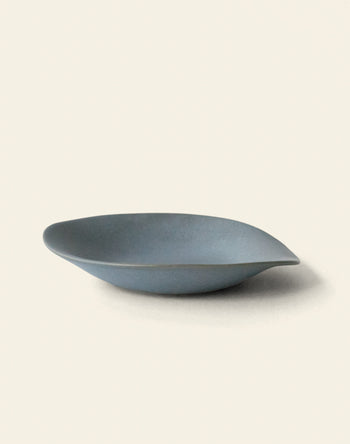 The NR Ceramics HIN Dessert Plate by NR Ceramics is a minimalist, matte blue-gray plate with a gently curved, organic shape, shown on an off-white background.