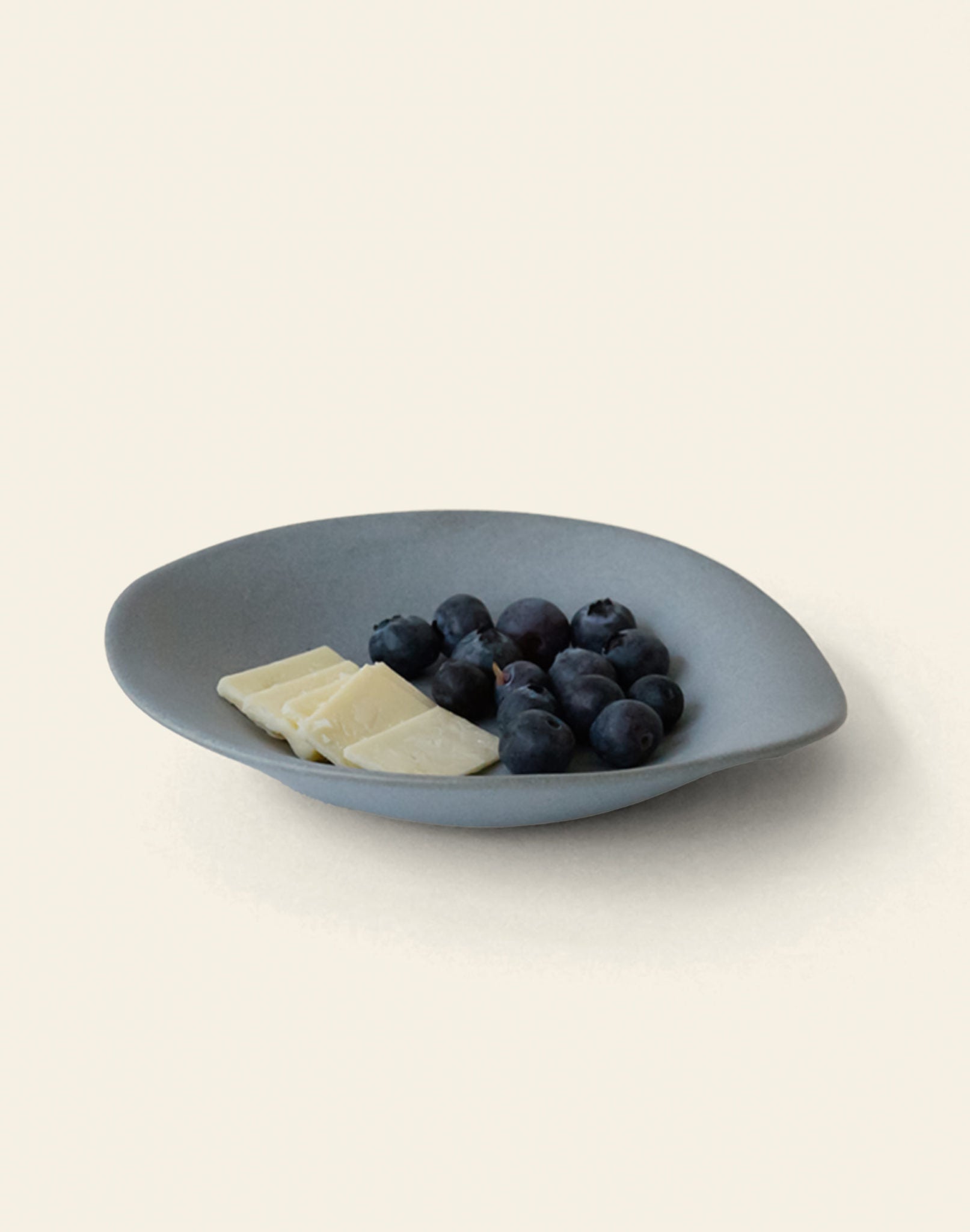 The NR Ceramics HIN Dessert Plate by NR Ceramics, in gray with an organic shape, displays fresh blueberries and three pieces of white chocolate on a simple light background.