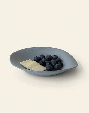 The NR Ceramics HIN Dessert Plate by NR Ceramics, in gray with an organic shape, displays fresh blueberries and three pieces of white chocolate on a simple light background.