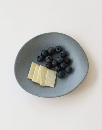 The NR Ceramics HIN Dessert Plate by NR Ceramics is shown on a white background with several fresh blueberries and four rectangular cheese slices arranged side by side.