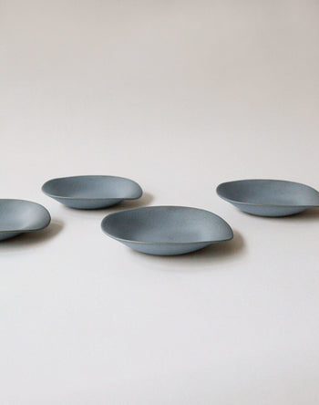 Four NR Ceramics HIN Dessert Plates, featuring a minimalist oval design, are arranged on a plain light gray surface to highlight their organic shape and the generous space between each plate.