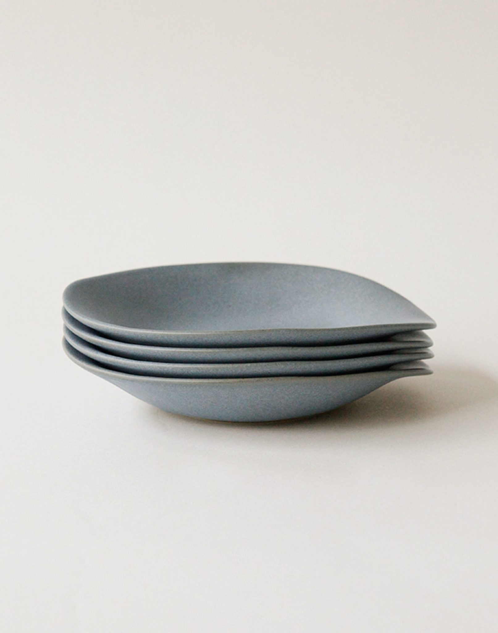 A stack of four NR Ceramics HIN Dessert Plates in matte gray, featuring an organic shape with slightly curved edges, is neatly arranged against a plain white background.