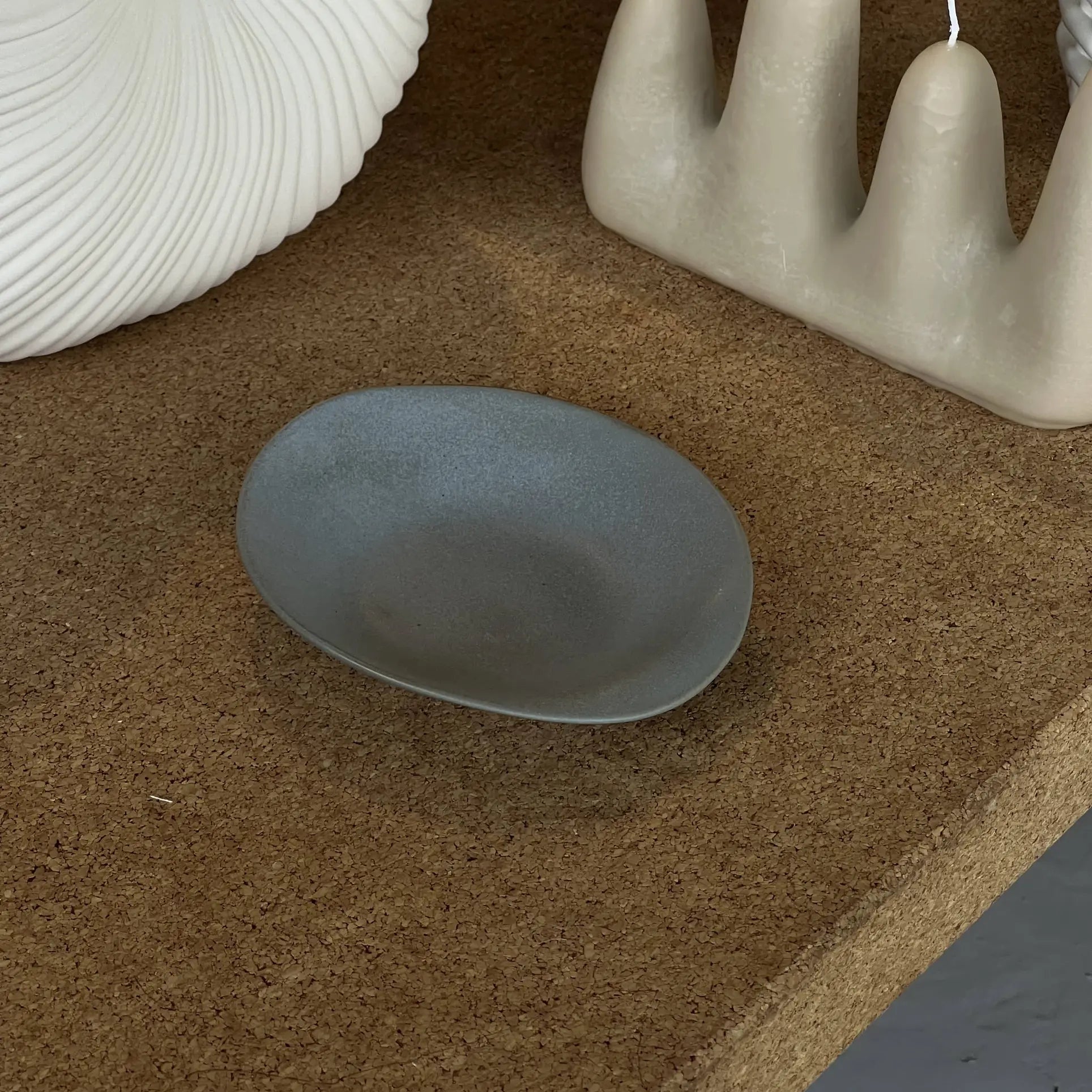 The NR Ceramics HIN Dessert Plate, a small matte gray ceramic piece by NR Ceramics, rests on a brown textured surface next to sculptural ceramic and wax decor in organic shapes and neutral tones.