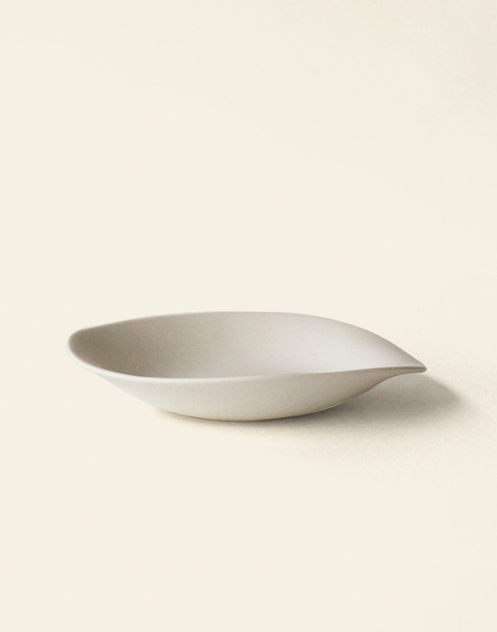 The NR Ceramics HIN Dessert Plate by NR Ceramics features a minimalist white ceramic design with an organic, leaf-inspired shape and smooth, curved edges, shown against a light neutral backdrop.