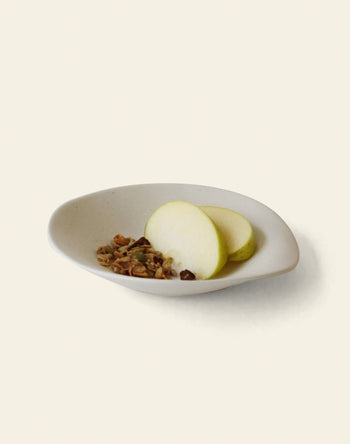 The NR Ceramics HIN Dessert Plate by NR Ceramics, with its organic shape in white, is shown on a light background holding two green apple slices and a small serving of granola.