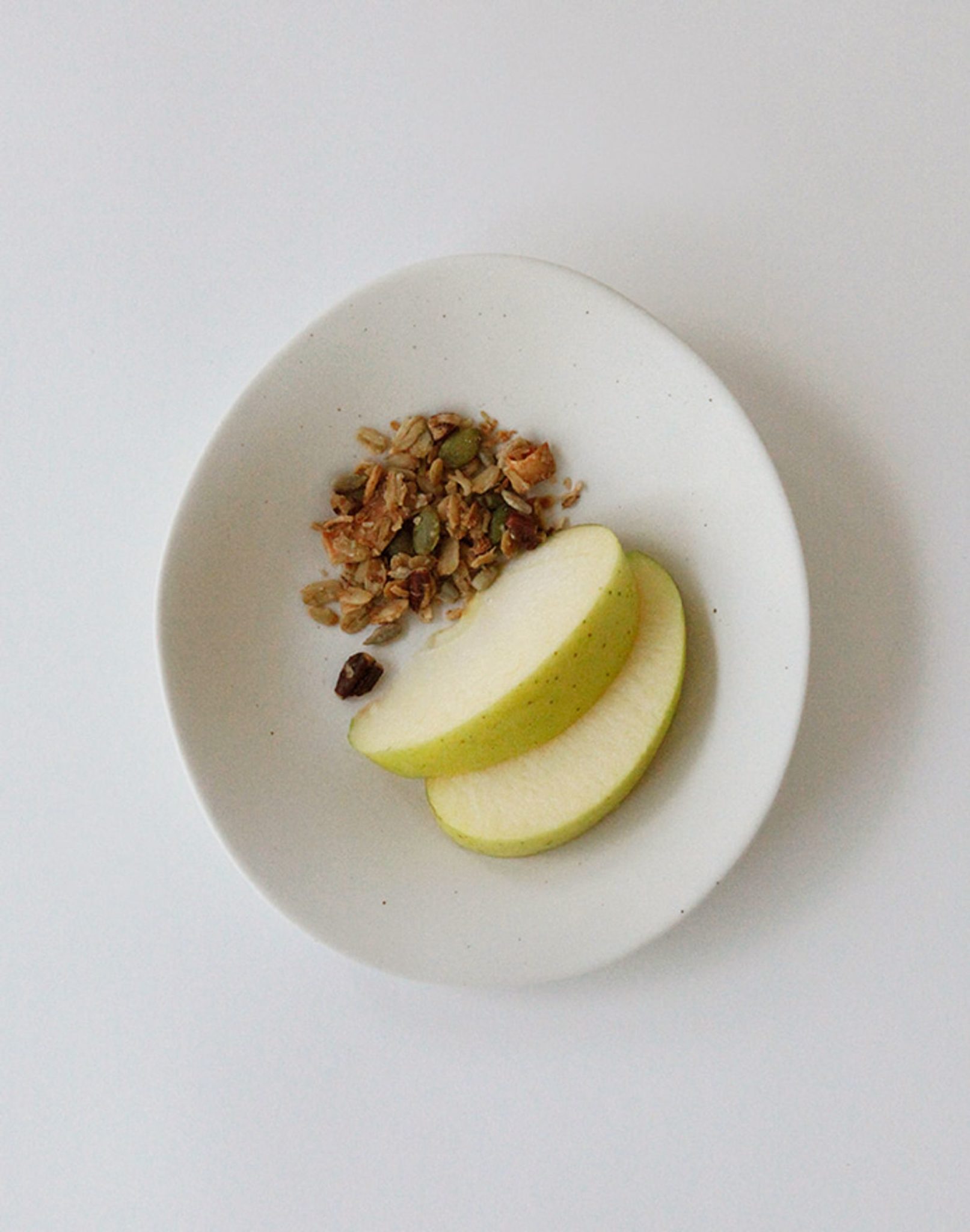 The NR Ceramics HIN Dessert Plate by NR Ceramics, featuring an organic white shape, holds two green apple slices and a small pile of granola on a plain white background.