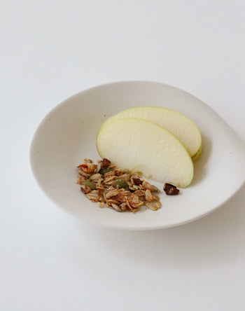 The NR Ceramics HIN Dessert Plate by NR Ceramics displays two green apple slices and a serving of seed-and-nut granola, arranged organically on a plain white background.