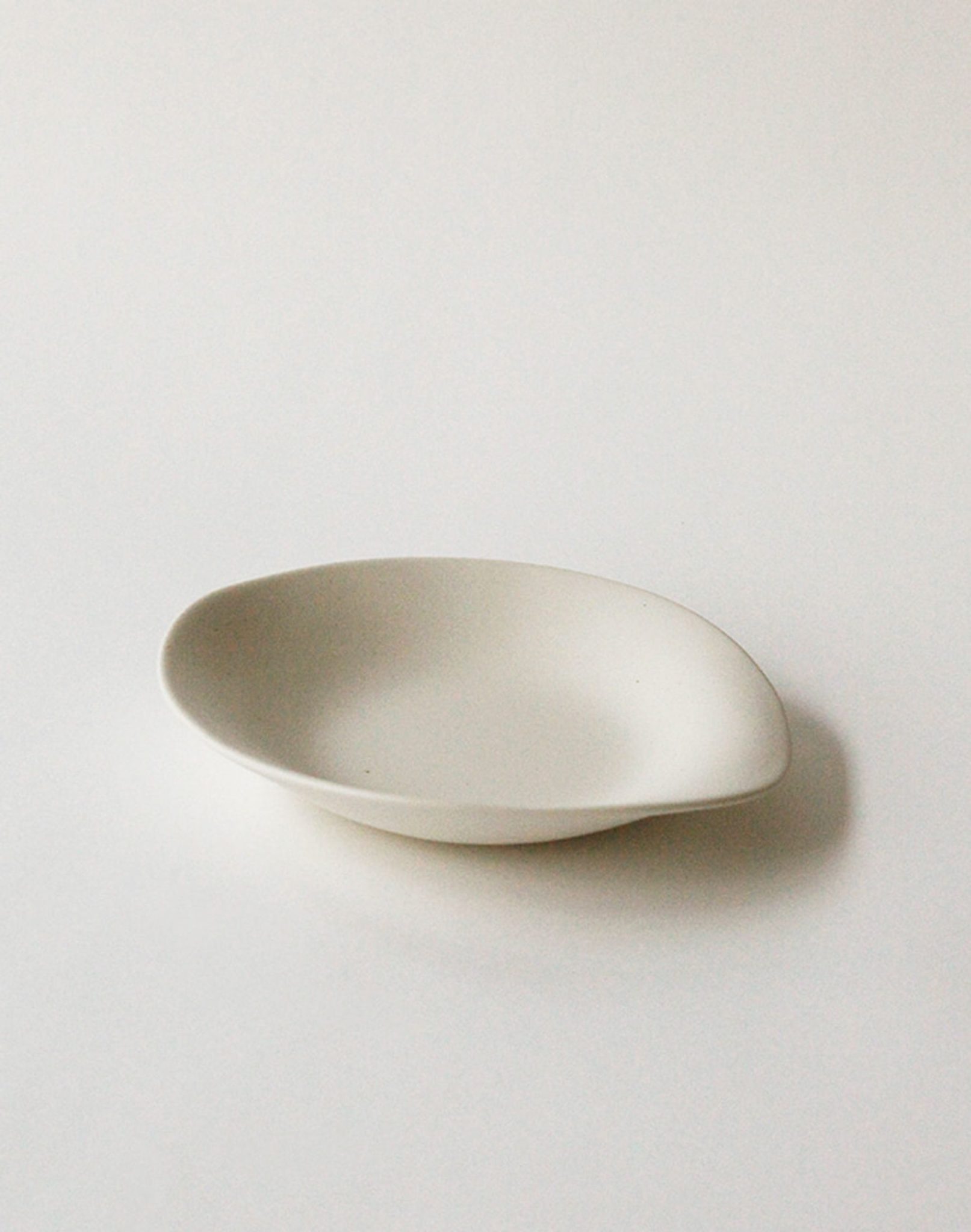 The NR Ceramics HIN Dessert Plate by NR Ceramics features an off-white finish and a slightly irregular oval shape. This plain, shallow plate from the Hin collection is shown resting on a white surface.