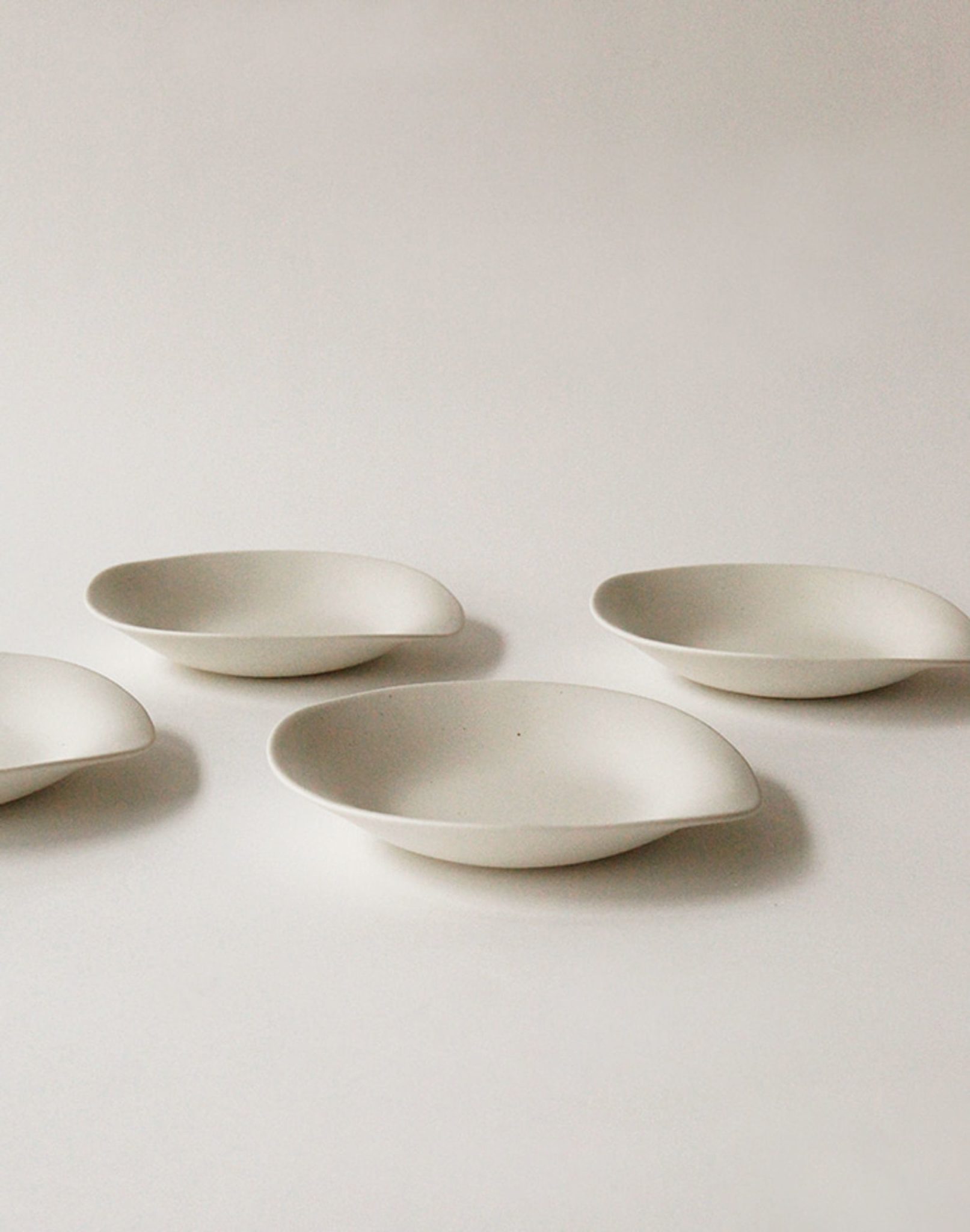 Four NR Ceramics HIN Dessert Plates are arranged on a plain white surface, evenly spaced with soft shadows that accentuate their minimalist design, modern aesthetic, and organic, oval-shaped white ceramic form.