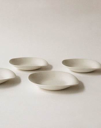 Four NR Ceramics HIN Dessert Plates are arranged on a plain white surface, evenly spaced with soft shadows that accentuate their minimalist design, modern aesthetic, and organic, oval-shaped white ceramic form.