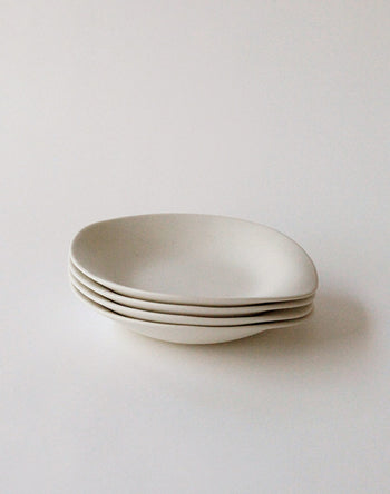 Four minimalist NR Ceramics HIN Dessert Plates in white ceramic are stacked on a light surface. Their subtle, organic shapes cast gentle shadows, highlighting the refined design by NR Ceramics.