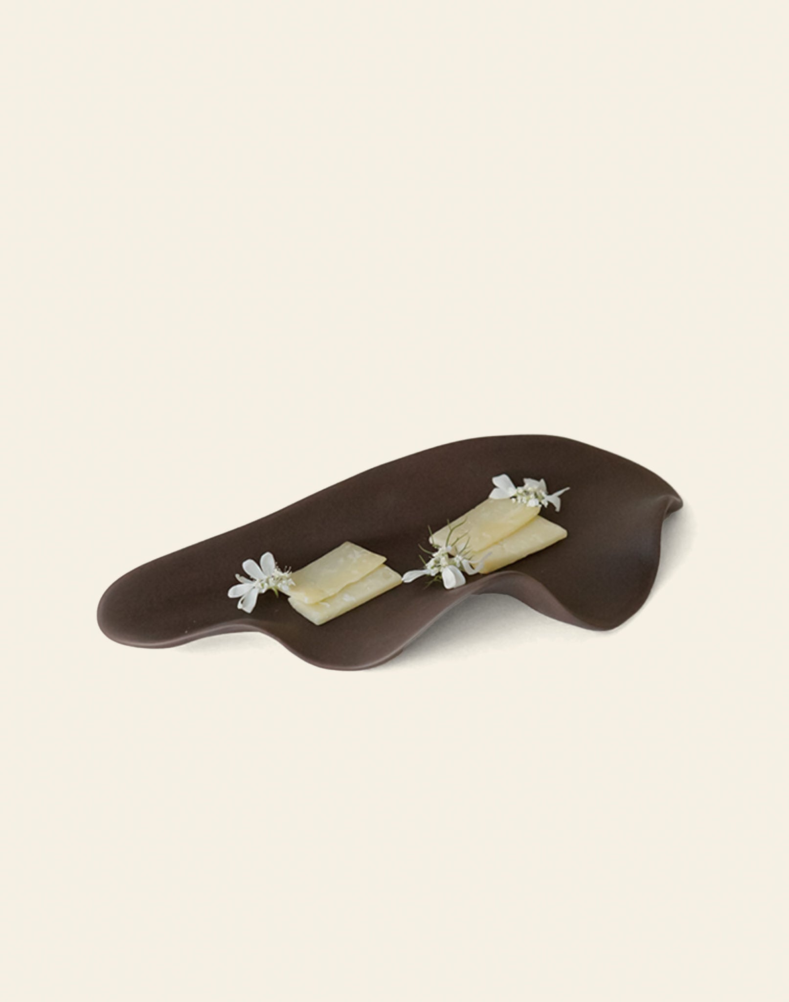 The NR Ceramics HIN Leaf Plate from NR Ceramics displays two rectangular cheese slices, topped with edible white flowers and herb sprigs, highlighting its multi-functional tableware design against a plain light backdrop.