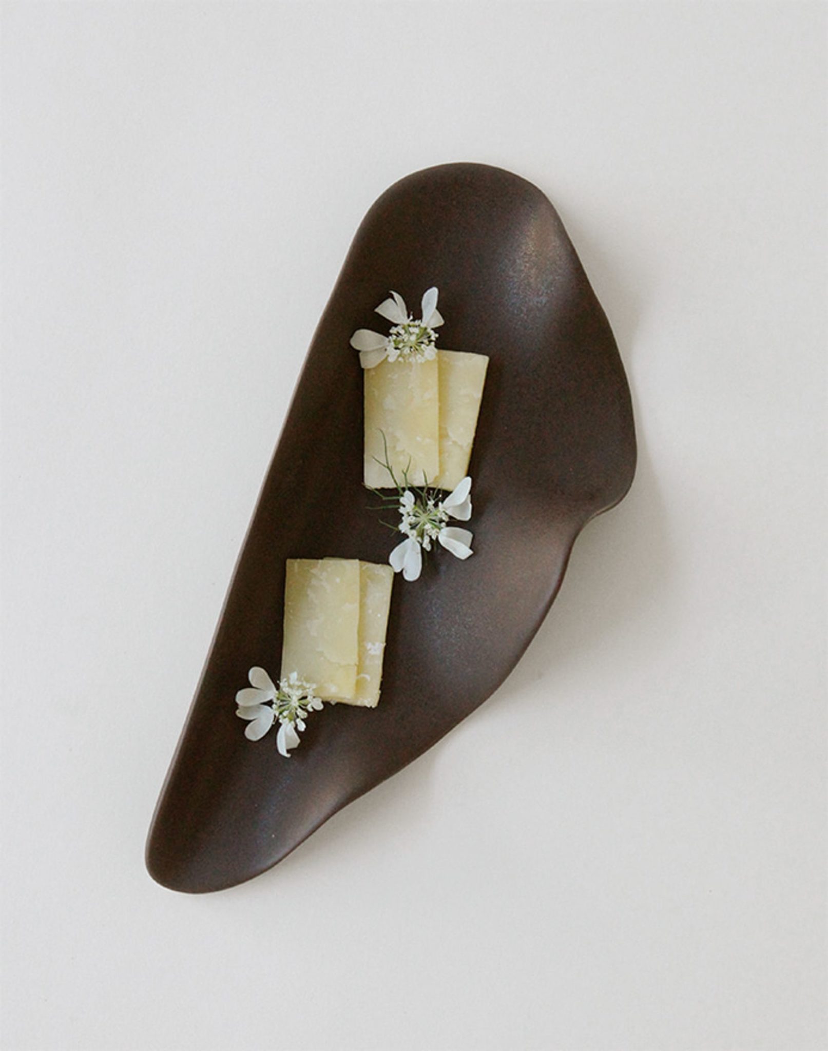 The NR Ceramics HIN Leaf Plate by NR Ceramics displays two small rectangular cheese slices topped with delicate white edible flowers, creating a striking contrast against a plain white background.