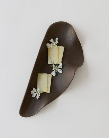 The NR Ceramics HIN Leaf Plate by NR Ceramics displays two small rectangular cheese slices topped with delicate white edible flowers, creating a striking contrast against a plain white background.