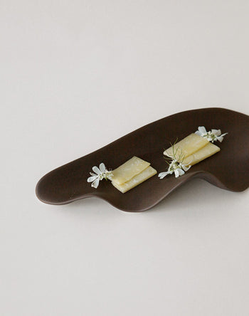 The NR Ceramics HIN Leaf Plate by NR Ceramics, in dark brown with a wavy design, elegantly displays cheese garnished with white flowers and herbs, highlighting its stylish and versatile use as tabletop serveware.