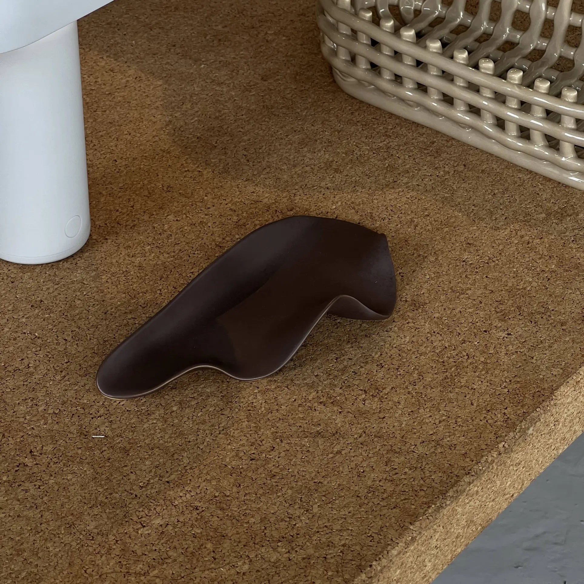 The NR Ceramics HIN Leaf Plate, a small, dark brown, irregularly shaped dish, rests on a cork surface next to multi-functional tableware—a woven basket and a white cylindrical container from NR Ceramics.