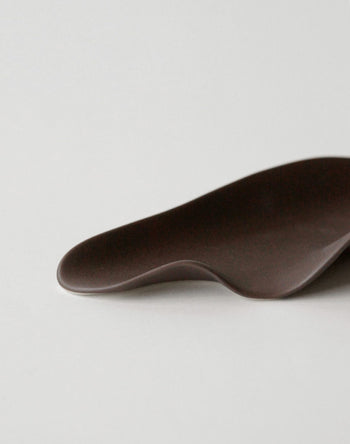 The NR Ceramics HIN Leaf Plate by NR Ceramics, featuring a dark brown abstract leaf shape with smooth curves, serves as elegant multi-functional tableware and is set against a plain light gray background.