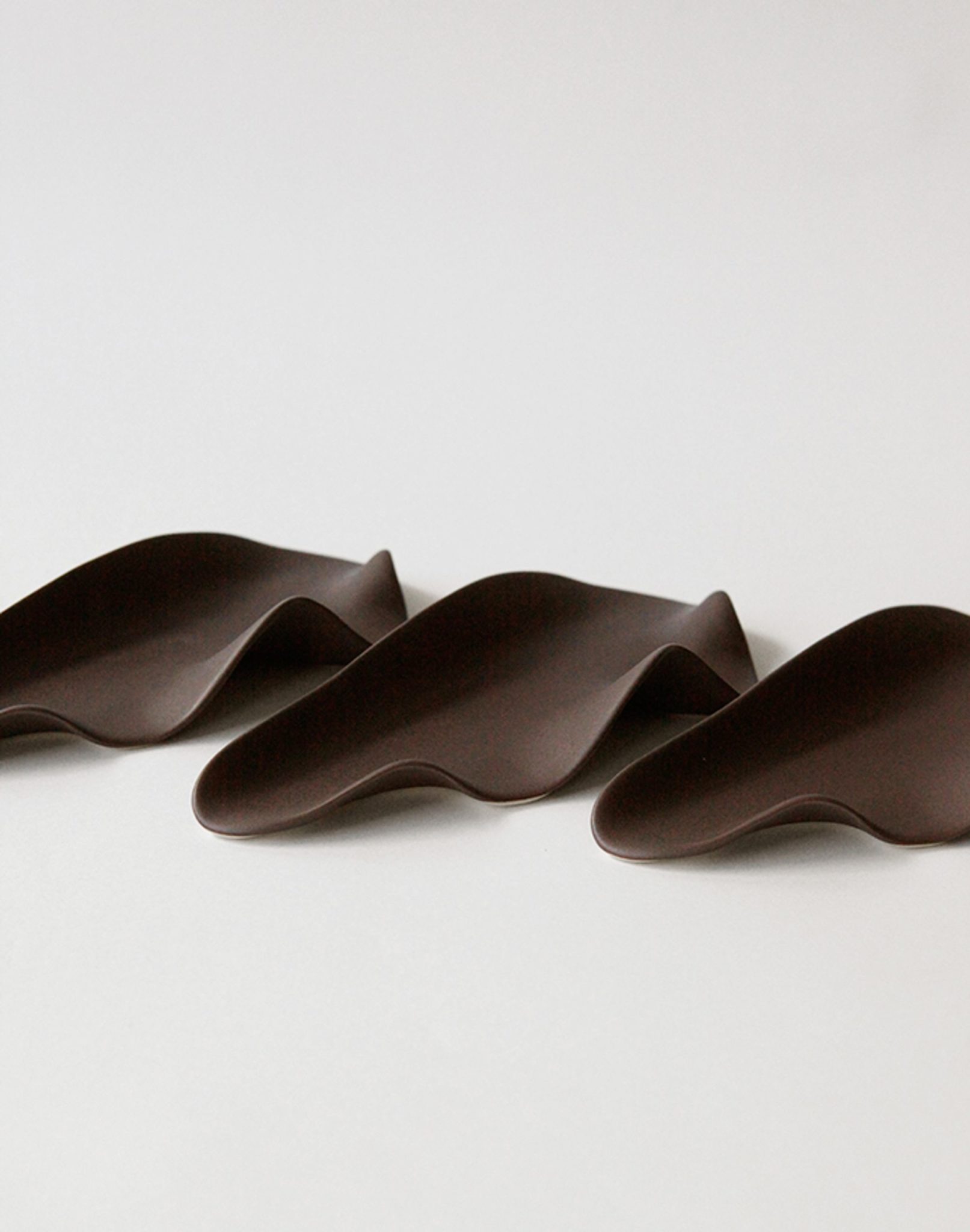 Three smooth, dark brown, wavy sculptural objects by NR Ceramics are arranged side by side on a light background—these multi-functional pieces echo the elegance of the NR Ceramics HIN Leaf Plate in contemporary tableware.