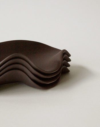 A stack of NR Ceramics HIN Leaf Plates, featuring dark, wavy edges, is arranged on a plain light surface with the overlapping plates forming a layered, sculptural look.