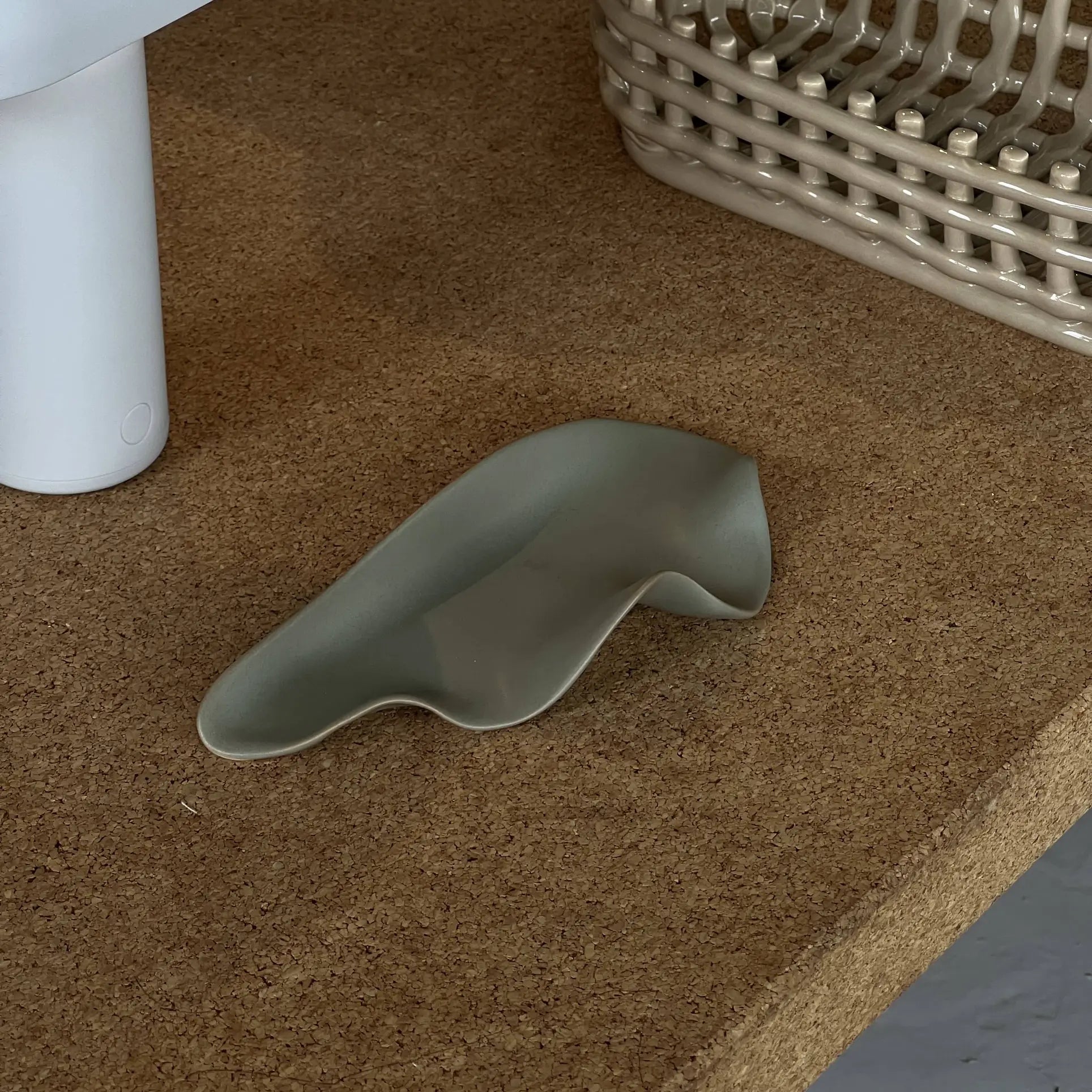 The NR Ceramics HIN Leaf Plate, a small, irregularly shaped green piece by NR Ceramics, rests on a cork surface beside a white cylindrical object and a woven basket.