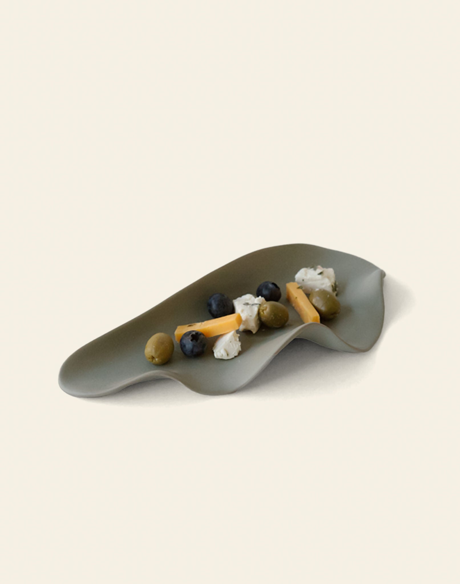 A stylish NR Ceramics HIN Leaf Plate in gray, by NR Ceramics, holds blueberries, green olives, cheese sticks, and soft white cheese—displaying its multi-functional use on a plain light background.