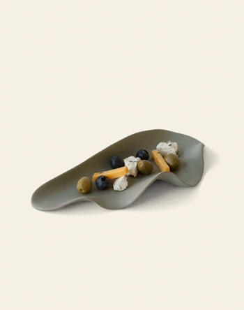 A stylish NR Ceramics HIN Leaf Plate in gray, by NR Ceramics, holds blueberries, green olives, cheese sticks, and soft white cheese—displaying its multi-functional use on a plain light background.