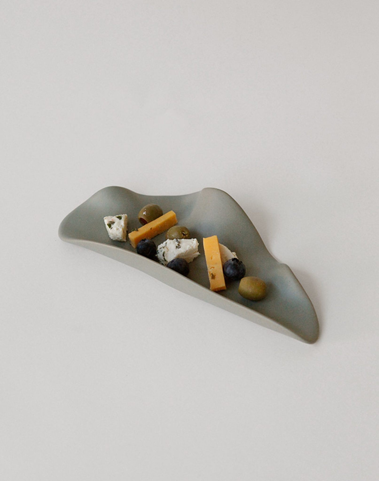 The NR Ceramics HIN Leaf Plate by NR Ceramics is a minimalist gray ceramic piece with an organic leaf shape, displayed as multi-functional tableware holding cheese, olives, blueberries, and a walnut on a plain white background.