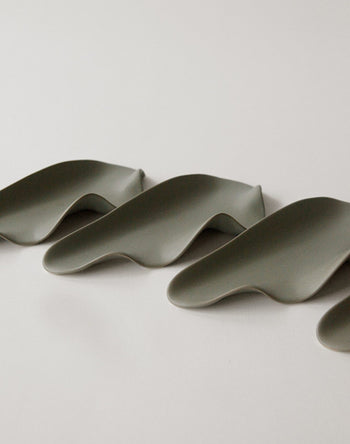 Four matte gray NR Ceramics HIN Leaf Plates from NR Ceramics are arranged in a row on a plain, light surface, showcasing their multifunctional, organically flowing design inspired by gentle undulating waves.