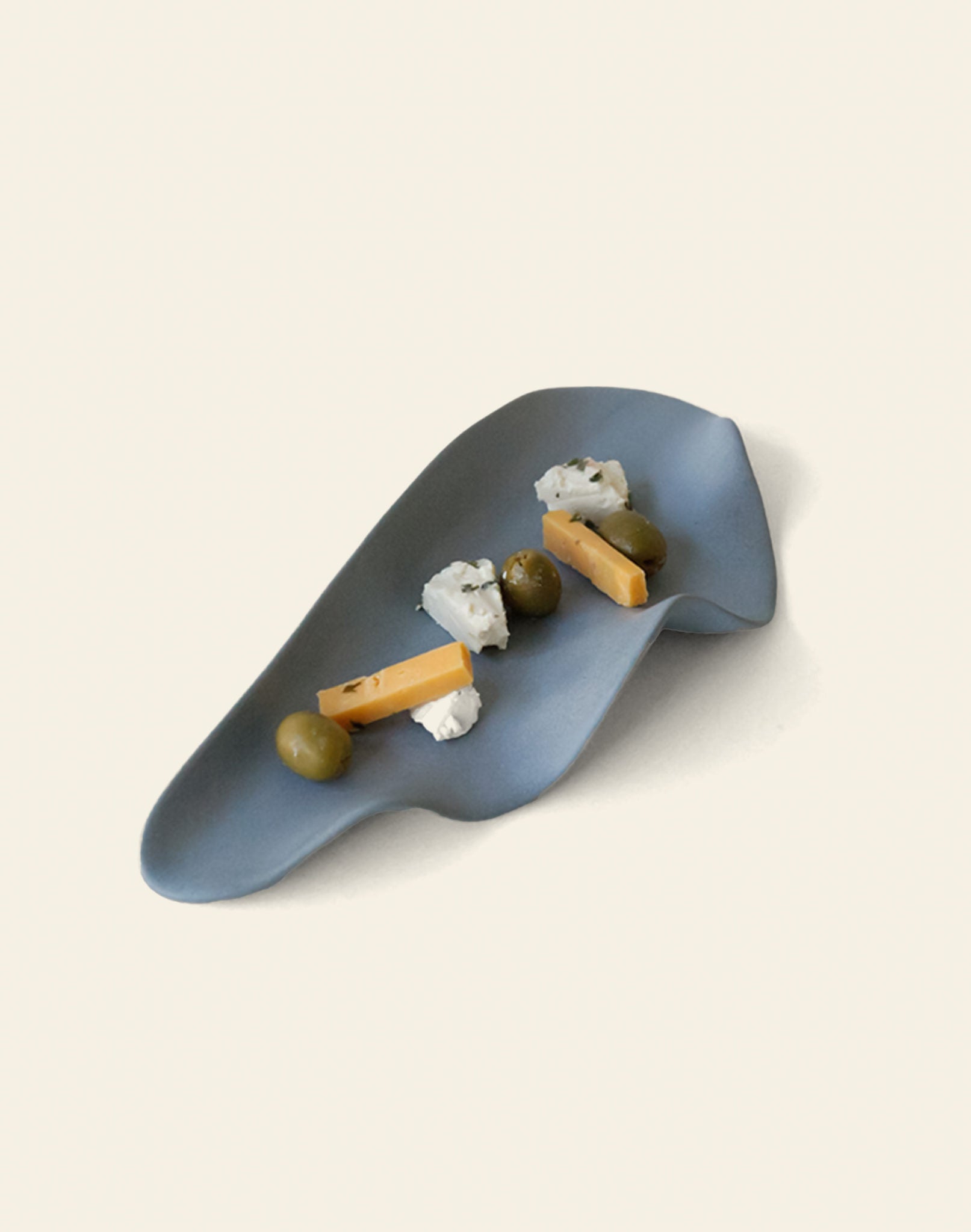 The NR Ceramics HIN Leaf Plate, in blue with an abstract shape, stylishly holds green olives, cheese sticks, and crumbly white cheese—perfect multi-functional tableware for elegant serving from NR Ceramics.