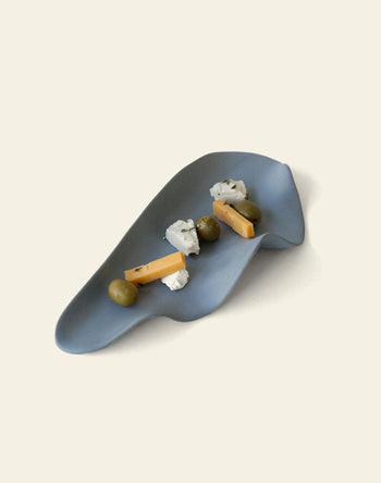 The NR Ceramics HIN Leaf Plate, in blue with an abstract shape, stylishly holds green olives, cheese sticks, and crumbly white cheese—perfect multi-functional tableware for elegant serving from NR Ceramics.