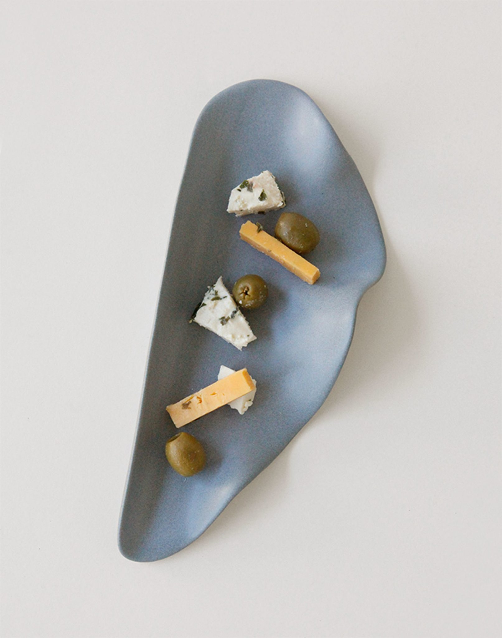 The NR Ceramics HIN Leaf Plate by NR Ceramics, featuring blue wavy edges, elegantly displays two white and orange cheese cubes and three green olives in a neat diagonal line—combining style with multi-functionality.